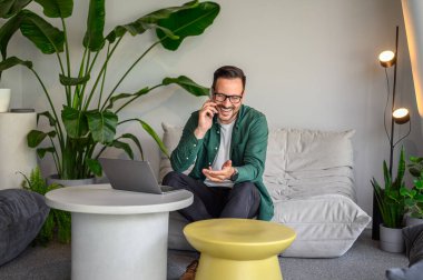 Modern işyerindeki koltukta otururken müşteriyle telefon görüşmesi yapmaktan mutlu bir genç.