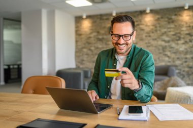 Genç yakışıklı girişimci ofisteki bilgisayardan kredi kartıyla online ödeme yapıyor.