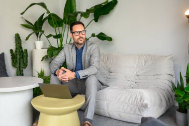 Ofisteki kanepede dizüstü bilgisayar başında çalışırken gözlüklü düşünceli genç işadamı gözlerini kaçırıyor.