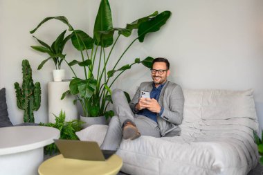 Modern ofiste kanepede otururken akıllı telefondan gülümseyen ve mesajlaşan yakışıklı bir işletmeci.