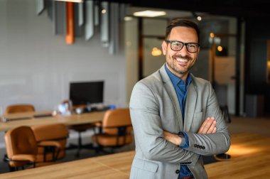 Kolları çapraz ve gözlükleriyle modern iş yerinde duran gülümseyen yakışıklı CEO portresi.