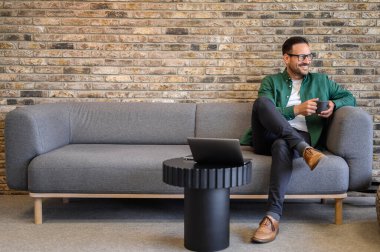 Ofisteki kanepede dinlenirken düşünceli bakışlarını kaçıran kahve fincanı olan pozitif erkek girişimci.