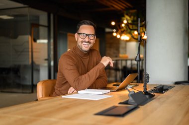 Gözlüklü, kameraya gülümseyen ve ofiste dizüstü bilgisayarla çalışan genç bir iş adamı.