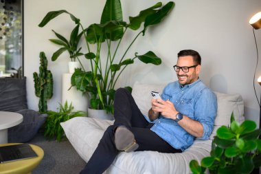 Modern işyerindeki kanepede dinlenirken cep telefonuyla mesajlaşan gözlüklü yakışıklı bir yönetici.
