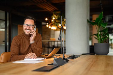 Erkek programcı, ofiste dizüstü bilgisayar başında çalışırken telefonda gülümsüyor ve projeyi tartışıyor.
