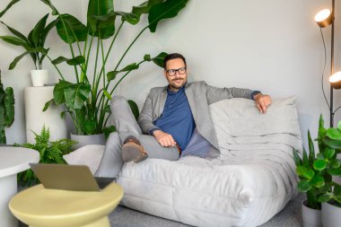 Modern işyerindeki kanepede rahat bir şekilde otururken gözlüklü düşünceli erkek CEO başka tarafa bakıyor.