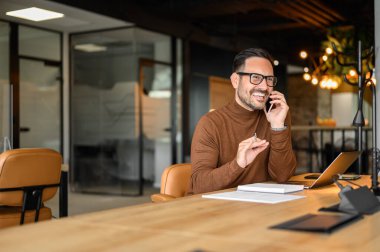Dizüstü bilgisayarı ve masasında not defteri olan neşeli profesyonel bir erkek iş yerinde akıllı telefondan konuşuyor.