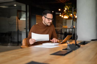 Gözlüklü, mali raporları ofisteki dizüstü bilgisayardan analiz eden odaklanmış bir muhasebeci.