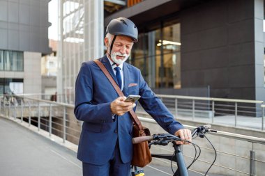 Kasklı erkek girişimci cep telefonuyla mesajlaşıyor ve şehirde yürürken bisikletini itiyor.