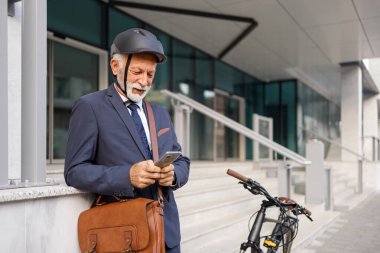 Çantalı ve kasklı yaşlı bir erkek yönetici bisikletle ofisin önünde dikilirken cep telefonu kullanıyor.