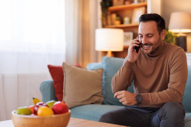 Oturma odasında kanepede otururken akıllı telefondan gülümseyen ve konuşan yakışıklı bir adamın portresi.