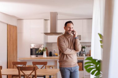Pencereden bakan, akıllı telefondan konuşan orta yaşlı bir adam yemek masasında beklerken gülümsüyor.