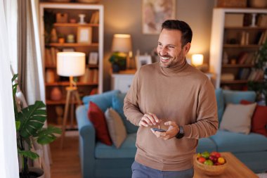 Yakışıklı, orta yaşlı bir adamın oturma odasında dikilirken gözlerini kaçırıp telefon kullanmasının portresi.