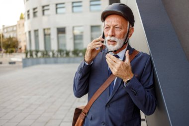 Kasklı ciddi bir iş adamının portresi. Şehirde duvarın yanında dururken akıllı telefondan dinliyor.