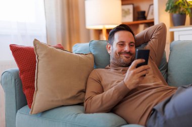 Yakışıklı adam evdeki oturma odasında kanepede dinlenirken akıllı telefondan gülümsüyor ve mesajlaşıyor.