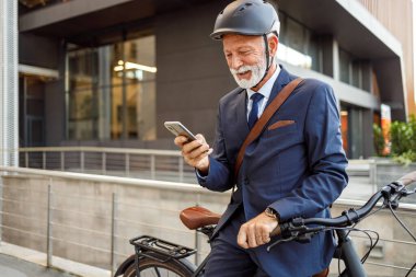 Kasklı ve bisikletli yaşlı sakallı işadamı işe giderken akıllı telefondan mesaj atıyor.