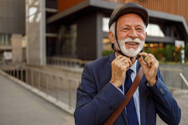 Bisiklet kaskı takan ve işe giderken kameraya gülümseyen kendinden emin yaşlı bir adamın portresi.