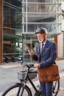 Kasklı yaşlı işadamı, bisikletle caddede dururken akıllı telefondan mesaj atıyor.