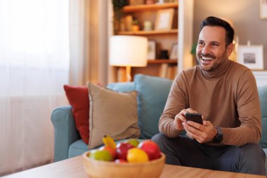 Neşeli orta yaşlı bir adam akıllı telefondan mesaj atıyor ve oturma odasında kanepede otururken gözlerini kaçırıyor.