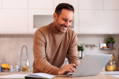 Modern mutfaktaki masada gülümseyen ve bilgisayarla çalışan yakışıklı girişimci portresi