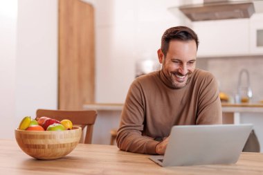 Yemek masasında otururken bilgisayarla gülümseyen ve çalışan yakışıklı serbest çalışanın portresi.