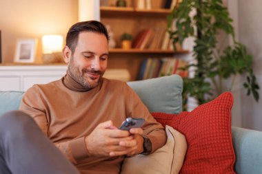 Evde kanepede otururken akıllı telefondan kendine güvenen erkek serbest mesajlaşmasının portresi.