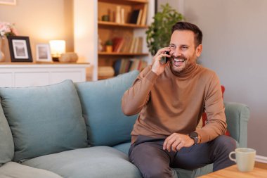 Ofisteki kanepede otururken akıllı telefondan konuşan neşeli orta yaşlı iş adamının portresi.