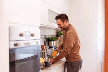 Gülümseyen orta yaşlı bir adamın modern mutfakta ev işi yaparken kirli bulaşıkları lavaboda yıkaması.