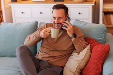 Yakışıklı bir girişimcinin portresi. Kahve içiyor, akıllı telefondan konuşuyor ve evdeki kanepede dinleniyor.