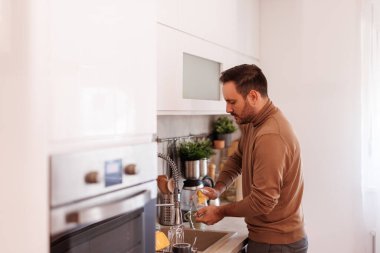 Ciddi orta yaşlı bir adamın mutfakta dururken lavaboda süngerle bardakları yıkamasının yan görüntüsü.