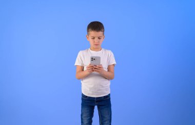 Mavi arka planda dikilirken akıllı telefondan mesajlaşan ciddi, sevimli bir çocuğun portresi.