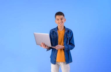 Dizüstü bilgisayar tutan ve mavi arka planda dikilirken kameraya gülümseyen kendinden emin genç bir çocuğun portresi.
