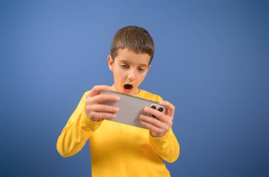 Odaklanmış, ağzı açık, mavi arka planda cep telefonuyla video oyunu oynayan genç bir çocuğun portresi