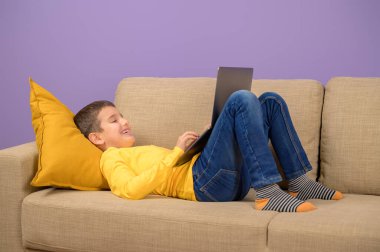 Kaygısız çocuk, pembe arka planda kanepede yatarken dizüstü bilgisayarda gülümsüyor ve oyun oynuyor.