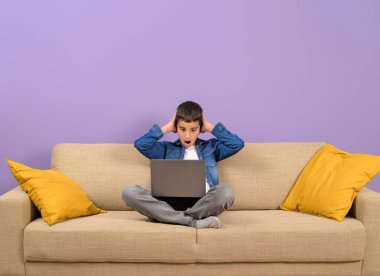 Kanepede mor arka planda otururken dizüstü bilgisayarından e-posta okuyan şaşkın çocuk.