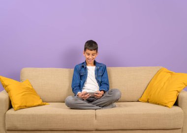 Gülümseyen genç çocuk, mor arka planda rahat bir şekilde koltukta otururken mobil oyunlar oynuyor.