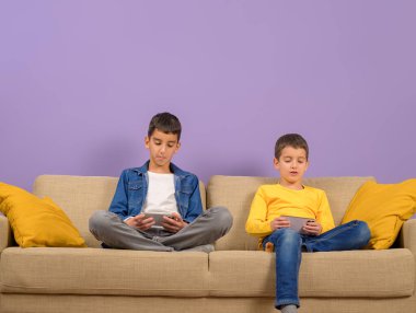 Mor arka planda kanepede yan yana otururken çevrimiçi oyun oynayan sevimli genç kardeşler.