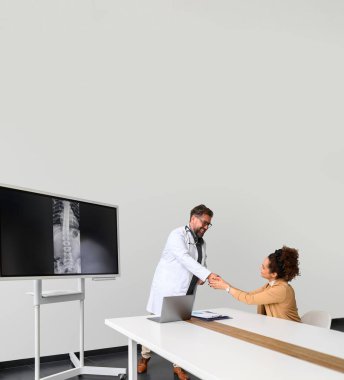 Kadın hasta hastanede masasında otururken el sıkışıyor ve erkek doktora şükranlarını sunuyor.