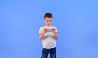 İzole edilmiş mavi arka planda dururken cep telefonuyla video oyunu oynayan çocuğun portresi