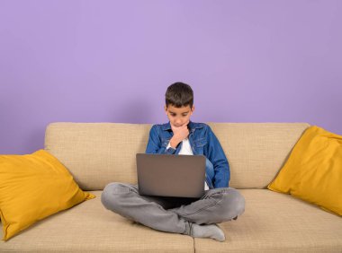 Mor arka planda kanepede otururken laptoptan bilgi okuyan düşünceli bir çocuk.