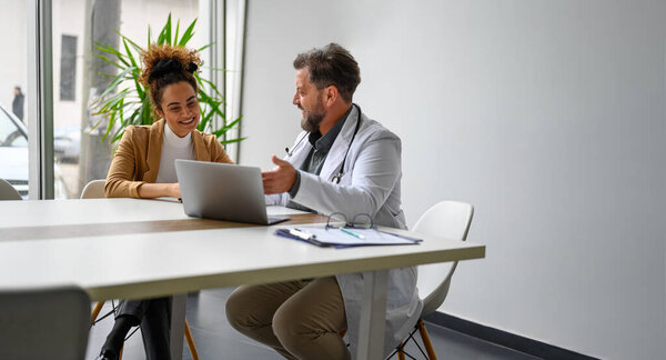 Врач-мужчина объясняет лечение или терапию с помощью ноутбука улыбающейся пациентке за столом в больнице