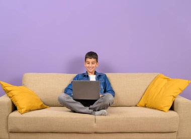 Gülümseyen genç işadamı, mor arka planda koltukta otururken dizüstü bilgisayardan veri analiz ediyor.