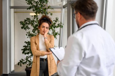 Endişeli genç kadın, erkek doktora semptomlarını açıklıyor. Hastanedeki not panosuna not alıyor.