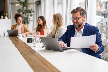 Mali belgeleri inceleyen ve arka planda kadın profesyoneller olan bir dizüstü bilgisayar kullanan erkek muhasebeci.