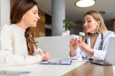 Kadın doktor elinde reçeteli ilaçlar tutuyor ve hastane masasındaki hastayla ilaç detaylarını tartışıyor.