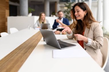 Mutlu yönetici, arka planda iş birliği yapan meslektaşlarıyla çevrimiçi toplantı sırasında proje detaylarını dizüstü bilgisayardan tartışır.