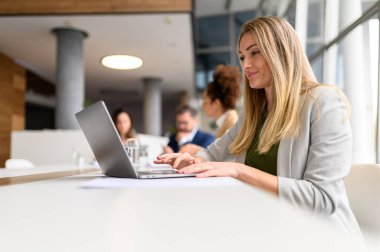 Odaklanmış sarışın iş kadını laptoptan veri analiz ederken aynı zamanda çalışma odasının arka planında iş birliği yapıyor.