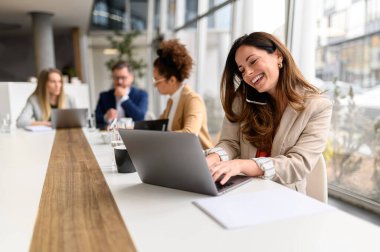 Sırıtkan avukat cep telefonuyla konuşuyor ve arka planda işbirliği yapan iş arkadaşlarıyla toplantı odasında dizüstü bilgisayarda e-posta yazıyor.