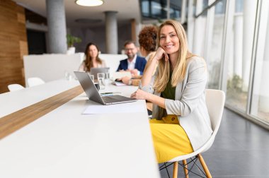 Masada dizüstü bilgisayarı olan sarışın yöneticinin portresi kameraya gülümserken, meslektaşlar da toplantı odasının arka planında iş birliği yapıyor.