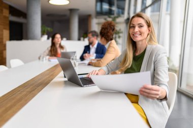 Rapor ve dizüstü bilgisayarla gülümseyen mali danışman arka planda profesyonel iş arkadaşlarıyla masada oturuyor.